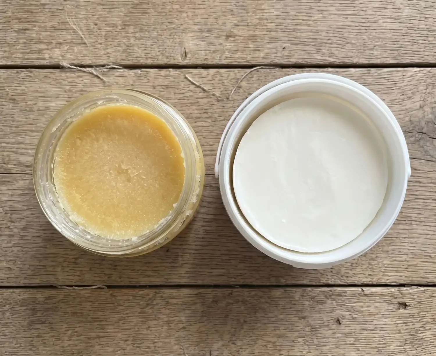 Top-down view of two open fat containers side by side on a rustic wooden surface — a glass jar of golden ghee on the left and a white tub of solid beef tallow on the right, showing the visual difference between the two cooking fats